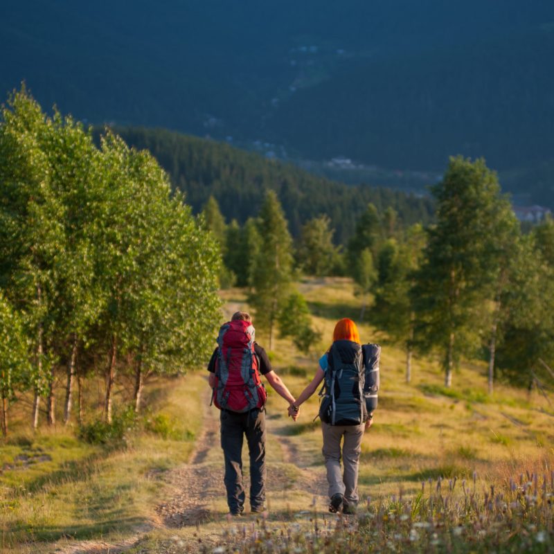 Back view couple hikers with backpacks walking along a beautiful mountain area and holding hands. Back view couple hikers with backpacks walking along a beautiful mountain area and holding hands. Lifestyle active vacations concept mountains landscape on background